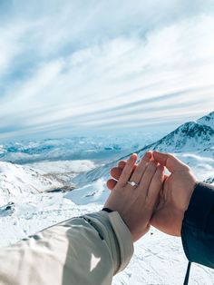 two people holding hands while standing on top of a snow covered slope with mountains in the background