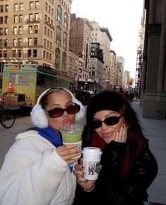 two women pose for the camera while holding drinks in front of them on a city street