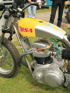 a yellow and silver dirt bike parked on top of a grass covered field next to people