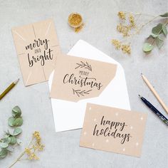 three christmas cards on top of a table next to some pens and pencils, with greenery