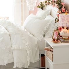 a white bed topped with lots of pillows next to a pink flower filled window sill