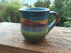 a blue and green mug sitting on top of a wooden table next to some bushes