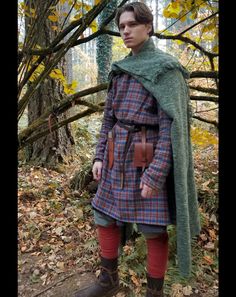 a young man dressed in medieval clothing standing under a tree with leaves on the ground