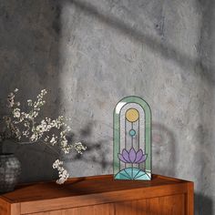 a vase with some flowers on top of a wooden table next to a wall and a stained glass window