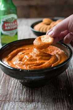 a hand dipping a cracker into a bowl of tomato dip with two bottles of beer in the background