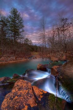 a stream flowing through a forest filled with rocks and trees under a cloudy sky at sunset