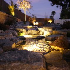 a garden with rocks and lights in the night time, along side a path that leads to a pond