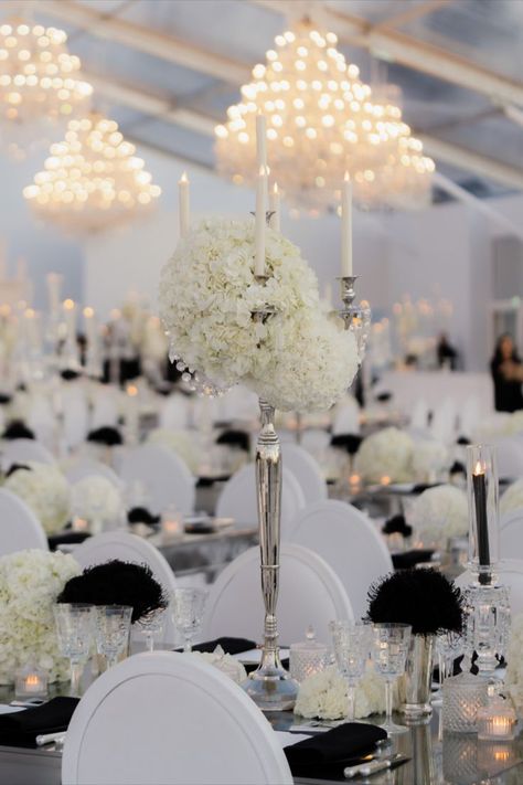 Immerse in luxury with this glamorous black and white wedding reception. Featuring elegant white hydrangea centerpieces, silver candelabras, and dramatic black and white chairs, the setting exudes sophistication. Crystal chandeliers above add a sparkling touch, creating a truly opulent atmosphere. Perfect for a black-tie affair or anyone seeking elegant wedding inspiration. Wedding decor, black and white wedding, glamorous wedding, wedding reception decor, wedding centerpiece, wedding flowers Modern Black And White Wedding Decor, Candelabra With Flowers, Wedding Decor Black And White, Black And White Chairs, Wedding Decor Black, Black And White Wedding Reception, White Hydrangea Centerpieces, Hydrangea Centerpieces, Elegant Black And White Wedding