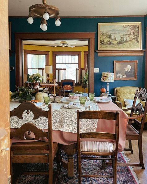 True vintage masterpieces are displayed in this deep blue dining room with thrifted decorations. From the retro green glasses to the mid-century light fixture, this eclectic design draws inspiration from every decade. Achieve a similar tablescape in your home by finding a delicate lace tablecloth, old dishes, and a little bit of pumpkin spice to inspire your holiday. Colorful Vintage Dining Room, Grandma Dining Room, Whimsical Dining Room, Vintage Thanksgiving Table, Farmhouse Thanksgiving Table, Vintage Dining Room Decor, Dining Room With Fireplace, Retro Dining Room, Old Dishes