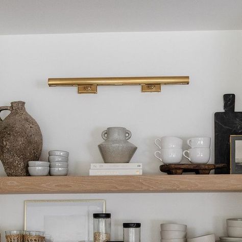 Visual Comfort & Co. on Instagram: "Shelfie appreciation post. 😍 Designer @rachaelbialkowski of @thelifestyledco⁣⁣⁣⁣⁠ helped her client @suzannavilla create a fresh yet timeless kitchen using a mix of neutral millwork and organic elements. Selected by the homeowner, the "cookies and cream" backsplash and countertop make a bold statement while pairing gorgeously with the white oak shelves for a natural look. Overhead, our iconic 30" Cabinet Maker's Picture Light by Chapman & Myers adds classic appeal to the space in a radiant Hand-Rubbed Antique Brass finish. The fixture brings a soft glow to the thoughtful collection of entertaining necessities and artful objects below. ​Architect: @harrisondesign 📷: @elizabethlaurengranger" Gallery Kitchen Lighting Ideas, Picture Light Kitchen Shelf, Picture Light In Kitchen, Cream Backsplash, Kitchen Bookshelf, Kitchen Built Ins, Lamp Kitchen, Bookshelf Lighting, Library Shelves