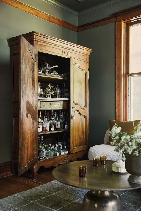 Dark and moody, the music room is coated in Benjamin Moore’s Army Green. It’s the perfect place to mix a drink at the antique armoire-cum-bar cabinet and linger around the vintage brass coffee table, both from Chairish. Armoire Bar Repurposed, Antique Bar Cabinet, Armoire Coffee Bar, Vintage Bar Cabinet, Armoire Ideas, Tall Bar Cabinet, Victorian Home Renovation, French Bar, Armoire Bar