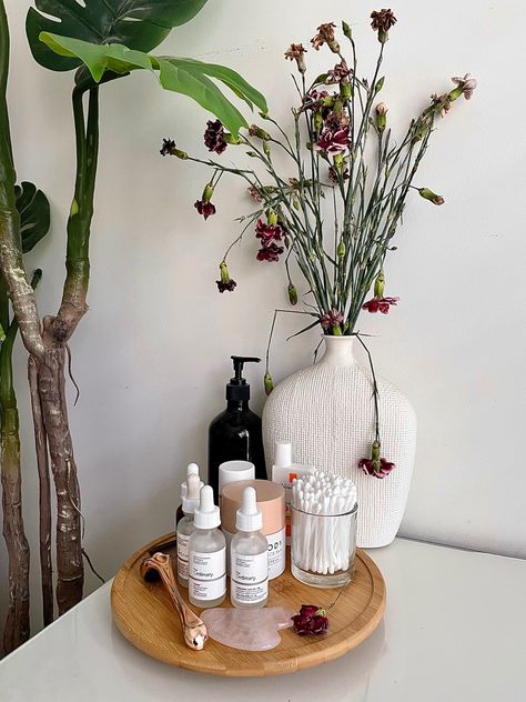 On a clean white desk there are magenta Flowers in a white vase, in-front of it is a wooden turntable with skincare products on it. Skin Care Desk Organization, Minimalist Skincare Organization, Skin Care Stand, Bathroom Skincare Display, Skincare Display Ideas, Flowers In Bathroom, Skincare Desk, Skincare Wall, Bathroom Skincare Organization