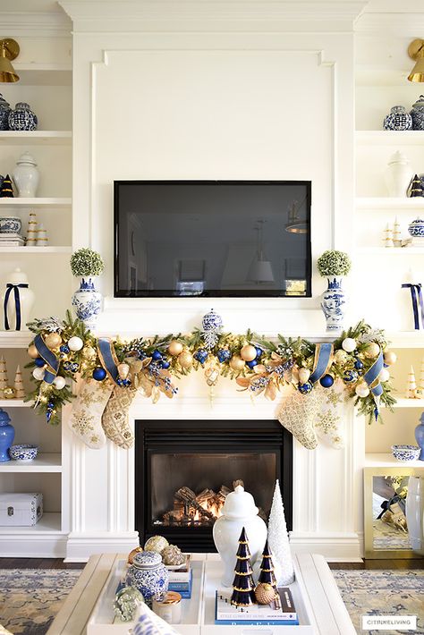 Christmas mantel decorated with faux green garlands, blue and gold ornaments and ribbon with gold and ivory beaded stockings. Blue and white chinoiserie vases with green topiary balls sit at each end of the garland on the mantel. Red And Gold Garland Christmas, Blue And Gold Christmas Decor, Chinoiserie Topiary, Gold Christmas Garland, Blue And Gold Christmas Tree, Ham Christmas, Grandmillennial Christmas, Christmas Mantel Ideas, Blue And Gold Christmas