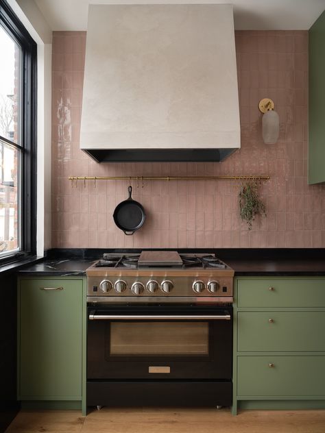 A beautiful green kitchen with tiles on the backsplash that are a taupe pink colour that pairs really well with the medium green of this kitchen. Along with all the brass accent with the handles, the rail, the sconce, the overall material palette of this project gives off a very unique and english vibe. The project is vibrant and colourful. We see big black windows that matches the dark soapstone countertop. Kitchens 2023, Green Kitchen Inspiration, Pink Kitchen Walls, Green Kitchens, Sage Kitchen, Pink Cabinets, Green Backsplash, Colourful Kitchen, Sage Green Kitchen