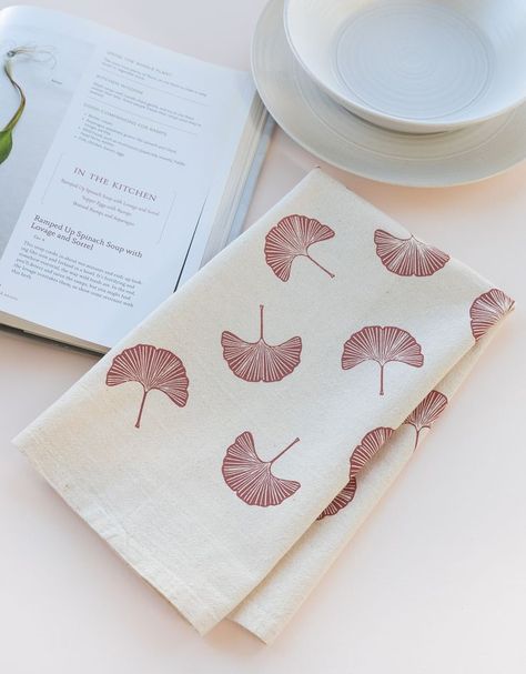 A natural cotton tea towel printed with a dusty rose ginkgo design is folded next to an open cookbook and a stack of dishes. Block Print Tea Towels, Ginkgo Leaf Pattern, Plant Kitchen, Hand Printed Textiles, Linocut Printmaking, Decorative Hand Towels, Printed Tea Towel, Ginkgo Leaf, Diy Crafts Hacks