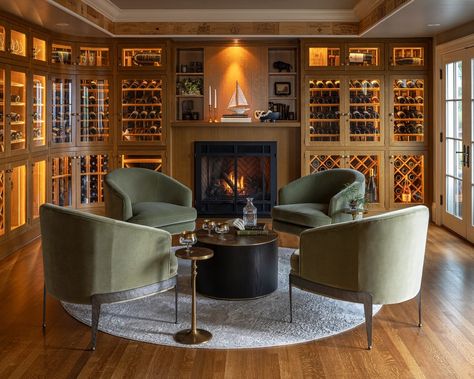 Elegant wine room with oak cabinetry featuring illuminated wine racks, four olive green velvet armchairs arranged in a circle, a round black coffee table, and a fireplace with a ship model on the mantle. Parlour Palm, European Interiors, Bar Lounge Room, Home Wine Bar, Parlor Room, Parlor Palm, Modern Farmhouse Home, Coffered Ceiling, Wine Room