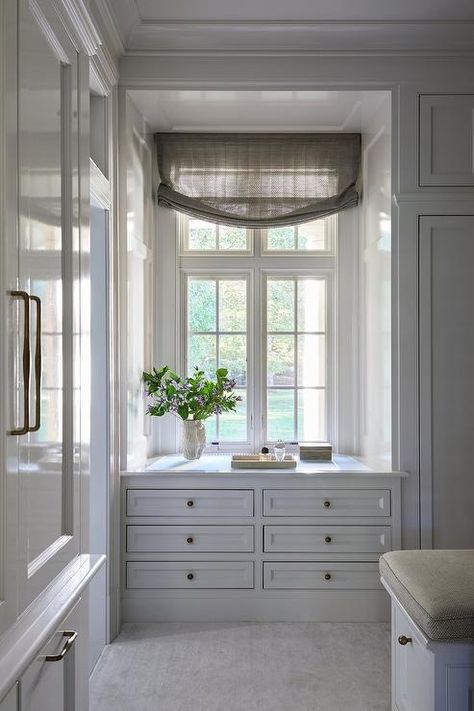 Chic walk in closet features a white built in dresser with brass knobs under a window with tan shade. Closet Between Windows, Built In Dresser Under Window, Pass Through Closet To Bathroom, Closet Designs With Window, Built In Linen Closet Hallway, Chic Walk In Closet, Hallway Linen Closet Built Ins, Closet With Built In Dresser, Dresser Under Window