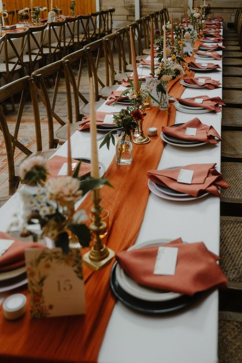 Wedding reception table decor with long burnt orange table runner and burnt orange napkins. Taper candles stand in brass candle holders & elegant Autumn flowers sit in small glass bud vases. Long trestle tables with cross back chairs. Wedding Table With Table Runner, Wedding Table Settings Burnt Orange, White And Burnt Orange Wedding Table, Burnt Orange Table Runner Wedding, Burnt Orange And Brown Wedding Decor, Rustic Wedding Decor Autumn, Diy Wedding Table Set Up, Autumn Wedding Diy Decorations, Wedding Reception Table Runners