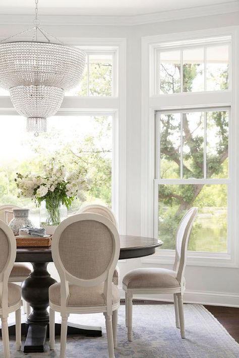 A black oval wooden dining table sits on a cream and blue rug surrounded by French round back ivory and gray dining chairs lit by an Aerin Jacqueline Two-Tier Chandelier. Bria Hammel Interiors, Bria Hammel, Round Back Dining Chairs, Cottage Dining Rooms, Woven Dining Chairs, French Dining Chairs, Transitional Dining Room, Echo Lake, Coastal Dining