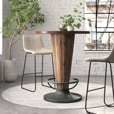 This round pub table really elevates your space with its edgy look and mix of industrial materials. It showcases a circular pedestal base made from metal with a weathered black finish and a solid wood body with an inverted cone shape that supports the table top. The visible grain variation and the saw marks add to its raw, salvaged look. And we’re pretty sure you’ll appreciate the ring around the base that doubles as a footrest. This table is ideal for smaller space, especially if your stools or Rustic Bar Height Table, Bar Tables And Chairs, Round Bar Table And Stools, High Bar Table Outdoor, Tall Circle Table Dining, Modern Bar Table Design, High Top Table Decor, Pub Table Decor Ideas, Tall Round Table