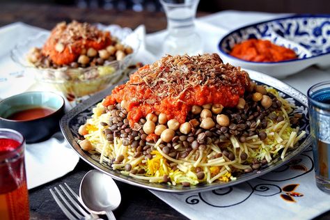 A plate of Koshari sits on a table set with smaller plates, cutlery, and middle easter inspired napkins Egyptian Recipes Authentic, Egyptian Dinner, Egyptian Koshari Recipe, Egypt Recipes, Koshari Recipe, Hummus Falafel, Egyptian Recipes, Egyptian Heritage, East Recipes