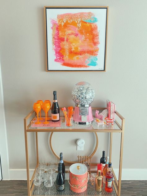 a cute gold bar cart with orange and pink accents. Funky Bar Cart Decor, Bar Cart Picture Frame, College Bar Cart Decor, Bar Cart Items, Bar Cart In Apartment, Decorative Bar Cart, Bar Cart Styling College, Pink Bar Cart Aesthetic, Bar Cart Simple