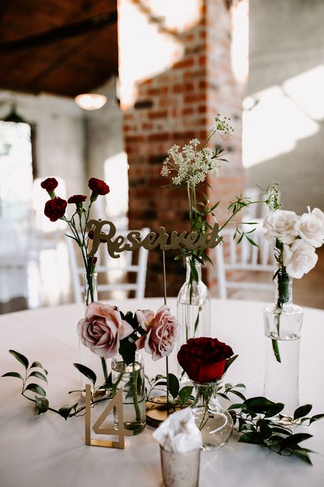 Wedding table setting with roses and other flowers in bud vases and table number. Bud Vases Wedding Centerpiece Winter, Burgundy Blush Centerpiece, Burgundy Bud Vase Centerpiece, Black And Burgundy Centerpieces, Burgundy Bud Vases, Moody Bud Vases, Single Flower Centerpiece, Wedding Centerpiece Burgundy, Vase Wedding Centerpieces