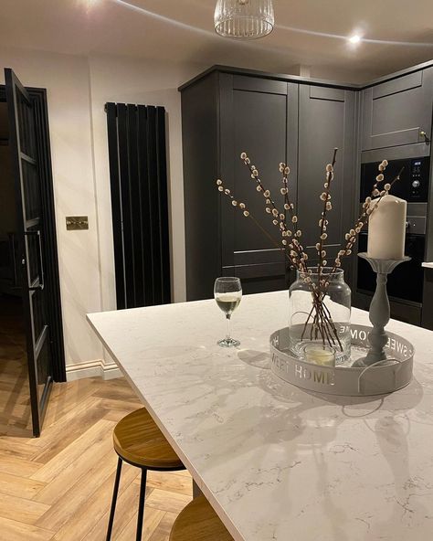 Light gray veins are spread across the white marble countertop in this modern farmhouse kitchen. The doors and wall structure match the dark gray cabinets that make a statement. Pair the table with wooden bar stools and a simple centerpiece of pussywillow branches in a glass vase to create a communal space. Dark Grey Kitchen Decor, Dark Gray And White Kitchen, Dark Gray Cabinets, Grey Kitchen Decor, Gray Kitchen Ideas, Modern Cottage Kitchen, Kitchen With Marble, Cool And Warm Tones, Simple Centerpiece