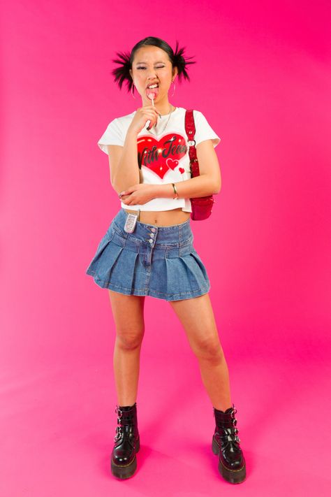 Girl standing in front of a pink backdrop licking a lollipop with her face scrunched up and one eye closed. She is wearing a white with jean top with a red heart on the front, pleated denim skirt, red vintage guess shoulder bag and black platform buckled doc martins. Early 2000s Editorial, Girly Aesthetic Photoshoot, 2000s Fashion Photography, Y2k Editorial Photoshoot, Y2k Fashion Editorial, Y2k Inspired Photoshoot, Y2k Fashion Photoshoot, 2000s Fashion Photoshoot, Early 2000 Photoshoot