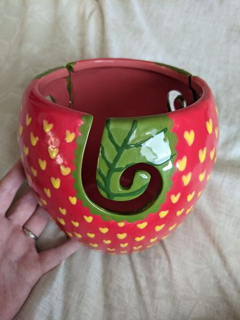 A red yarn bowl with yellow hearts painted all over it to look like a strawberry. The yarn bowl has a ceramic swirl yarn feeder that is painted green to mimic a leaf. A hand props up the bowl against a cream fabric backdrop. Yarn Bowl Pottery Ideas, Pottery Crochet Bowl, Cute Clay Yarn Bowl, Yarn Bowls Pottery Painting, Ceramic Yarn Bowl Painting Ideas, Painted Yarn Bowl, Yarn Bowls Pottery Diy, Polymer Clay Yarn Bowl, Knitting Bowls Pottery