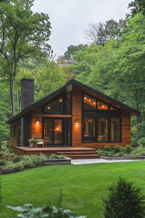 Modern cabin with a wooden deck surrounded by lush greenery. Uncover how sleek design, cozy woodwork, and a dash of minimalist charm are transforming cabins from rustic hideaways to luxurious retreats. Modern Log Cabin Floor Plans, Modern Cabin Big Windows, Woodsy House Plans, Cabins With Garage Underneath, Cozy Cottages In The Woods, Beautiful Forest House, Log Cabin Addition Ideas Exterior, Black Log Home Exterior, Small Home In The Woods