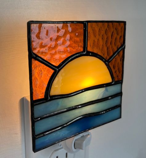 This 4 x 4 inch "Sun over Ocean" night light is made of stained glass using the copper foil or "Tiffany" style method. The colors are blue, amber and yellow opalescent glass. The seams are a polished black patina. The night light is attached to a UL listed night light switch module and includes a 4 watt bulb. Stained Glass Nightlight Patterns, Stained Glass Starry Night, Stained Glass Nightlight, Stained Glass Night Lights Patterns, Stained Glass Sun, Stained Glass Night Lights, Ocean At Night, Tiffany Stained Glass, Wall Plug