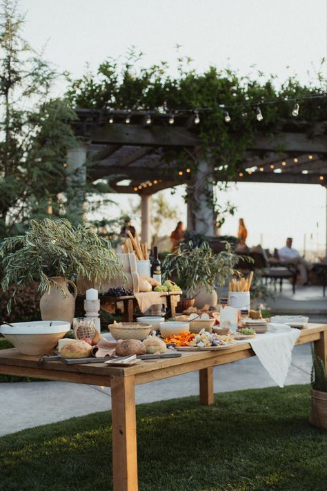 A photo of a large charcuterie table. It has large vases of olive branches and different breads. Bowls filled of seasonal fruits, hummus, and sliced bread. Looks like it is right out of the coast of Italy. Wedding Canape Table, Summer Wedding Buffet, Outdoor Grazing Table, Charcuterie Station Wedding, Appetizer Table Wedding, Garden Charcuterie Board, Charcuterie Board Wedding Reception, Engagement Party Food Table, Charcuterie Table Wedding