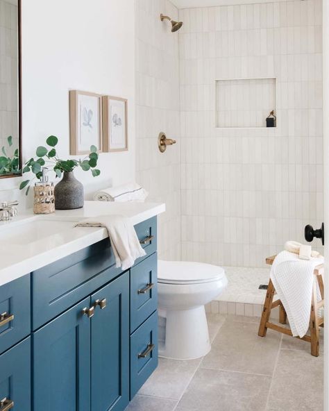 A door with a black knob stands open, revealing a blue cabinet, a white countertop, and a beige tiled floor. White towels on a wooden stool sit next to an off-white vertical accent tile in the shower. A gold faucet hangs over the gold shower handle. Blue Bathroom Ideas Decoration, Blue And White Bathroom Ideas, Bathroom Tiles Blue, Vertical Shower Tile, Bathroom Vanity Blue, White Bathroom Inspiration, Stone Shower Floor, Blue Bathroom Paint, White Shower Tile