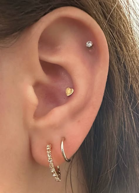 an ear with four piercings. two lobe piercings with the first being a small diamond hoop and the second being a smaller gold hoop. there is a gold heart conch piercing and a diamond stud high helix/cartilage piercing. Heart Conch Piercing, Conch And Mid Helix Piercing, Triple Lobe And Conch Piercing, Stud Conch Piercing, Helix And Lobe Piercing, Conch Stud Piercing, Helix Stud Piercing, Piercing Curation, Helix And Conch Piercing