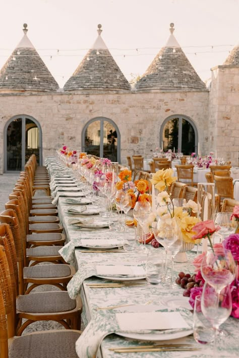 The wedding dinner was surrounded by trulli. Centerpieces evoke the Hindu theme with the eccentric color palette and the fruits spread on the tables. The flower arrangements are made in a modern style, with hand-worked roses. The green toile-de-jouy gives a beautiful touch, harmonizing all the colors in a surprising way. Wedding Centerpieces Aesthetic, Indian Dinner Table Decor, Italian Wedding Colour Schemes, Eclectic Wedding Theme, Green Toile Wedding, Pattern Tablecloth Wedding, Hindu Wedding Decor, Toile Wedding, Sicilian Wedding