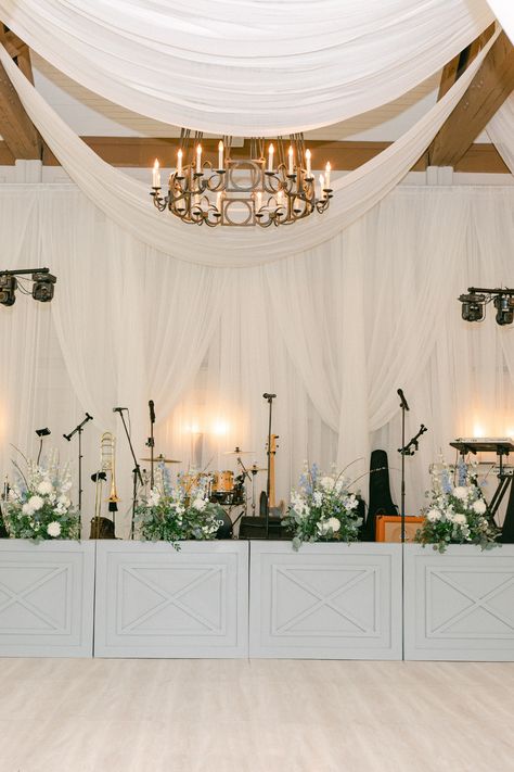 Ever detail of this wedding was covered including this gorgeous wedding stage. Assymetrical, wildflower style arrangements were used to continue the theme. The band backdrop was draped in white. #weddingdecor #weddingreception Stage Covering Wedding, Stage Florals Wedding, Wedding Band Stage Flowers, Wedding Band Stage Decor, Band Stage Decor, Stage Decor Wedding, Band Backdrop, Band Stage, Draping Wedding