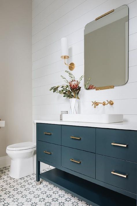 Fixed to a white shiplap wall, a frameless mirror with a brass trim is hung above a dark blue bath vanity accented with brass hardware and a vessel sink matched with a brushed gold cross handle wall-mount faucet. Teal Bathroom, Blue Vanity, White Shiplap Wall, White Bathroom Tiles, House Bathrooms, White Shiplap, Coastal Bathrooms, Transitional Bathroom, Bathroom Tile Designs