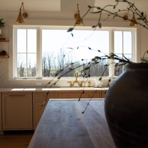 Three brass swing arm sconces are mounted over large kitchen windows. The marbled slab backsplash and brass faucet create a modern look when paired with neutral shaker cabinets sharing the same brass hardware. Window grilles are used to form sections, giving the large window a more homey appearance. Kitchen Windows Instead Of Upper Cabinets, Wide Window Kitchen, Kitchen Window Ledge Ideas, Large Kitchen Picture Window, Sink With Window Kitchen, Oven Between Two Windows, Lights Over Kitchen Window, Sconces Over Kitchen Window, Window Behind Kitchen Sink