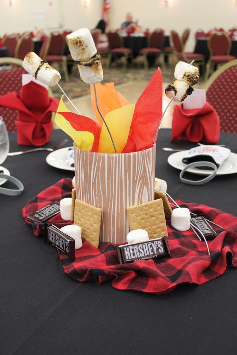 S’more and Camping Inspired Centerpieces -I used cardboard bird feeders (tops cut off) and Kleenex boxes from Michaels and wrapped with wood grain paper. Sand weighs down the box/gives placement to skewers. Dehydrate marshmallows prior. Put marshmallows onto skewer first and roast. Spray with matte finish spray paint which helps keep the ash from burning from falling off. Add some tissue paper for a fire affect and then place on top of sleeping bag like material with other S’mores-like goodies. Christmas Camping Theme, Camp Party Table Decor, Camping Theme Table Decorations, Montana Themed Party, Camp Theme Centerpieces, One Happy Camper First Birthday Centerpiece, Happy Trails Retirement Party, Camping Retirement Party Ideas, Campfire Centerpiece Ideas