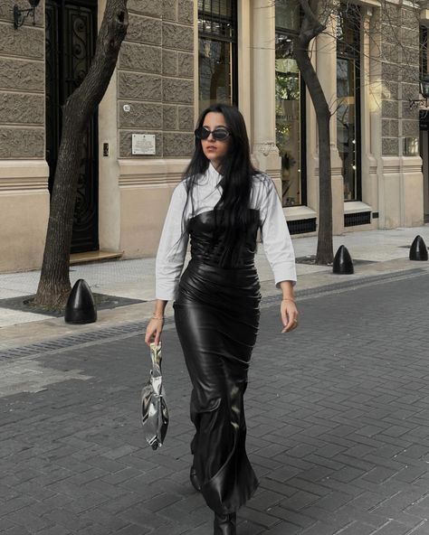 I am wearing a leather corset dress over a white shirt, accessorized with a metallic silver baguette bag and black sunglasses, posing by an elegant building facade.  Opción 2 White Shirt And Corset Outfit, Corset Winter Outfit, Corset And Shirt, Corset Street Style, Corset Over Shirt Outfits, Corset Over Shirt, Corset And Skirt Outfits, Leather Corset Outfit, Black Corset Outfit