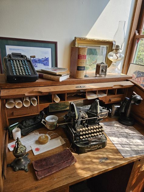 A black typewriter sits on a wooden desk next to an glass oil lamp, an old telephone, a teacup and maps. Rolled up paper, books and a framed painting sit on the top of the old desk. Light Academia Desk Setup, Roll Top Desk Decorating Ideas, Writer Desk Aesthetic, Light Academia Desk, Desk Aesthetic Dark Academia, Vintage Desk Ideas, Dark Academia Aesthetic Desk, Chaotic Academia Desk, Writing Desk Aesthetic