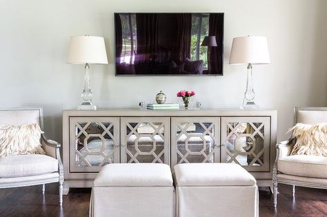 Chic living room features a pair of gray French chairs and oatmeal linen skirted ottomans placed in front of a gray mirrored cabinet fitted with mirrored trellis doors topped with crystal lamps placed under a flat panel tv. Wall Hanging Decorations, Mirror Wall Hanging, Under Tv, Crystal Lamps, Mirrored Sideboard, Dining Room Buffet, French Chairs, Transitional Living Rooms, Chic Living Room