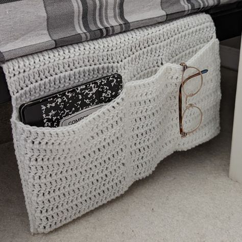 A white crocheted organizer hangs from the side of a bed. In its pockets are an ipad, a TV remote, and a pair of glasses. Crochet Book Nook Free Pattern, Bedside Organizer Crochet Pattern, Crochet Bedside Caddy, Crochet Bed Caddy, Crochet Book Organizer, Crochet Bed Pocket, Crochet Bed Organizer, Diy Bedside Organizer, Crochet Locker Organizer