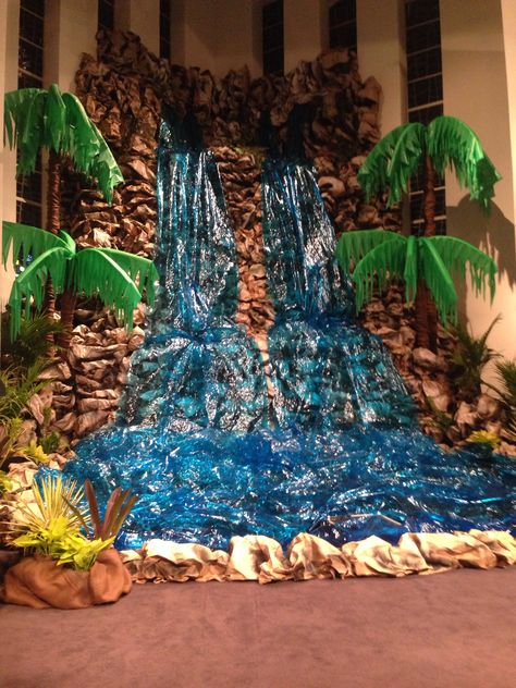 Waterfall made from kraft paper being stapled to cardboard and spray painted. Cellophane and lights for the water. Palm trees made out of carpet rolls and the leaves are cut pool noodles wrapped with table clothes: Fake Waterfall, Cave Quest Vbs, Cave Quest, Post Prom, Deco Jungle, Vbs Themes, Prom Decor, Football Decorations, Vbs Crafts