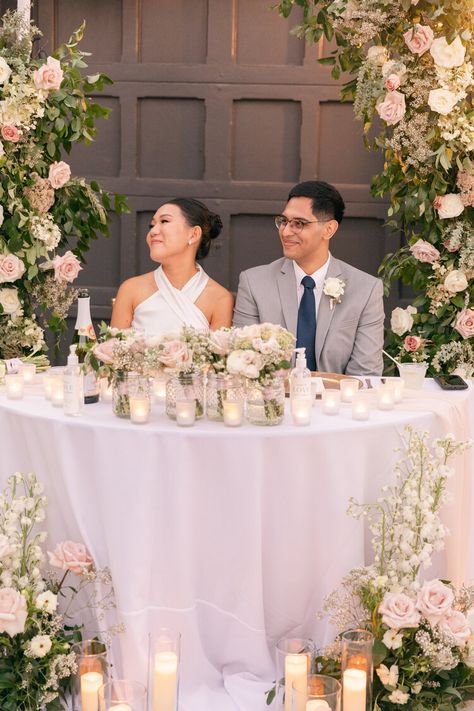 A sweetheart table full of blush floral decorations and candle details. The bride and groom look so happy sharing this moment together. Bride And Groom Table Arch, Sweetheart Table With Bridesmaid Bouquets, Wedding Bride And Groom Table Elegant, Bride And Groom Table In Front Of Fireplace, Half Circle Sweetheart Table, Burgundy Blush Sweetheart Table, Bridge And Groom Table, Sweetheart Table Bouquet, Bride And Groom Table Flower Arrangement