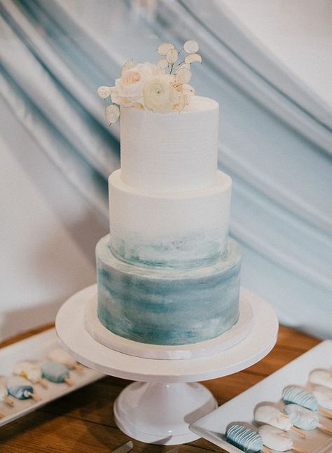 this three-tiered wedding cake is anything but boring with a plain white top and dusty blue ombre look at the bottom tier white roses are the decoration detail #weddingcake #weddinginspo Wedding Cakes Dusty Blue, Ems Cake, Wedding Cake Dusty Blue, White And Blue Wedding, Plain White Top, Wedding Cake Ombre, Beachy Wedding, Wedding Cake Roses, Ocean Wedding