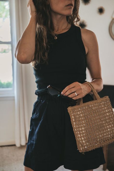 ashley wearing a black ribbed tank, black paperbag shorts, black strappy sandals, and a woven bag with tortoise shell sunglasses; a black mid century modern side chair in the background Black Belted Shorts Outfit, Minimal Chic Style Outfits Summer, Black Shorts Outfit 2023, Black Shirt Black Shorts Outfit, Black Tank Outfit Summer, Edgy Classic Summer Outfits, Black Ribbed Tank Top Outfit, Summer Outfit Black Skirt, Black Mom Shorts Outfit