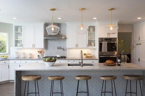 A white herringbone tiled island seats backless wood and metal stools at a honed white and gray waterfall countertop finished with a stainless steel sink and a satin nickel gooseneck faucet illuminated by three brass and glass schoolhouse pendants. Tiles On Kitchen Island, White Herringbone Tile, Green Kitchen Island, Kitchen Island Bench, Kitchen Island Decor, Kitchen Backsplash Designs, Stools For Kitchen Island, Fluted Glass, White Herringbone