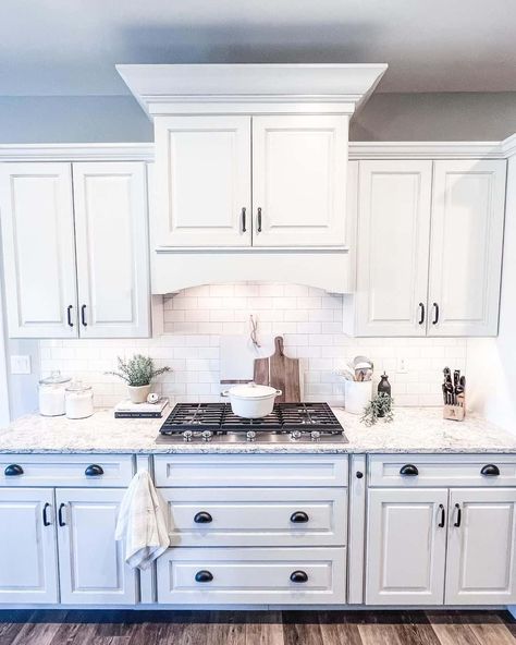 This modern farmhouse kitchen uses an arching cabinet casing with white paint to cover the range hood above a built-in gas stovetop. Glass apothecary jars, indoor succulents, cookbooks, and farmhouse utensil jars give the space a cozy vibe. Cabinet Range Hood Ideas, Range Hood Ideas, Farmhouse Kitchen Cabinet, Grey Marble Kitchen, Glass Upper Cabinets, Kitchen Range Hoods, Creative Cabinet, Shiplap Kitchen, Kitchen Eating Areas